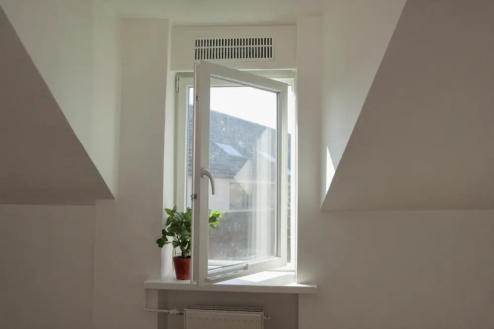Een openstaand raam in een dakkapel met een ventilatierooster erboven en een plant op de vensterbank