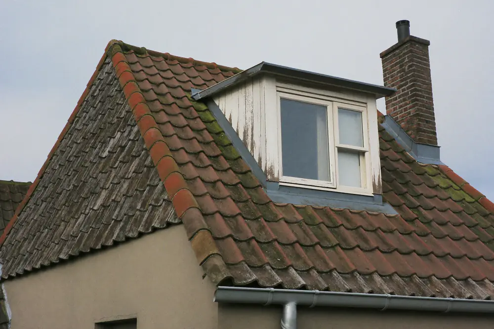 Oude houten dakkapel met afbladderende verf en verweerde boeiboorden op een jaren 70 woning